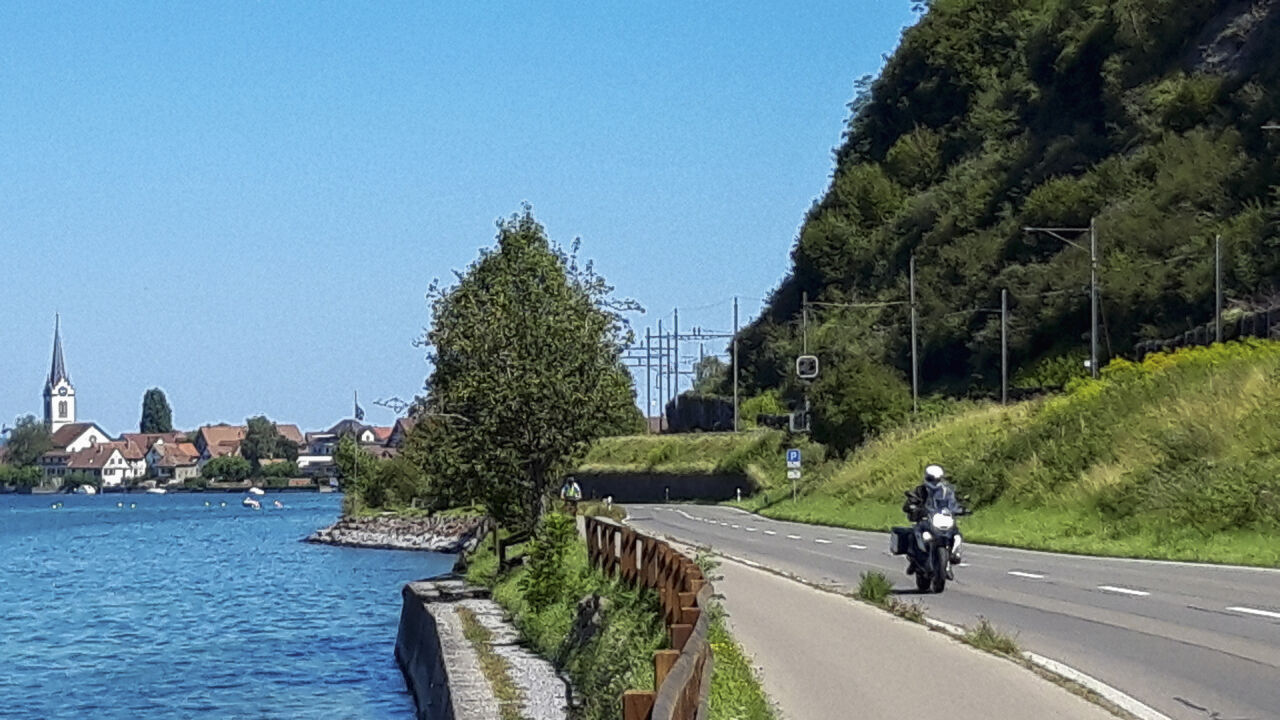 Motorrad auf einer Uferstraße, links der See, im Hintergrund ein Dorf mit Kirchturm
