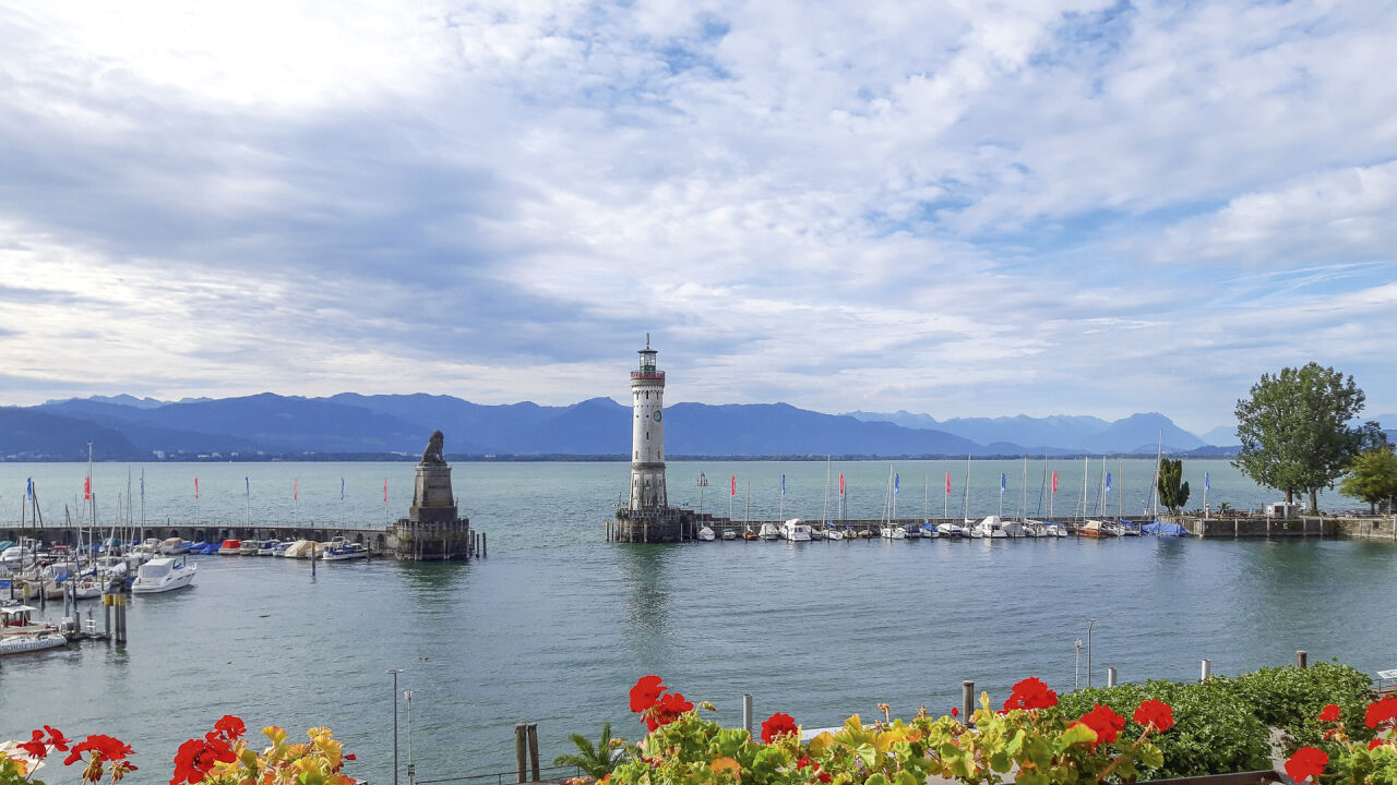Hafeneinfahrt mit Leuchtturm und Löwenstatue auf der Mole, dahinter Berge über dem See